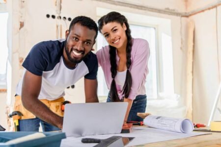 smiling couple using laptop and looking at camera while making renovation of home